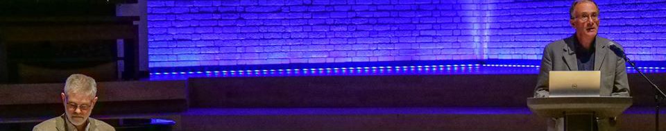 Man giving a speech on a stage with spotlights, whilst another man takes notes