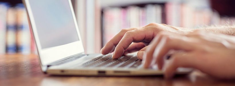 Hand typing on a laptop keyboard with bookshelf in the background