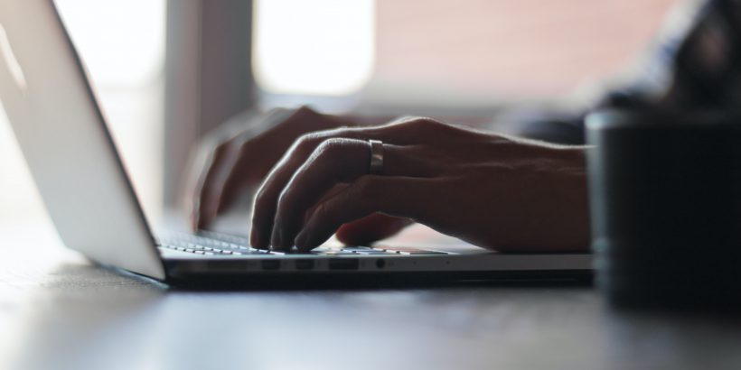 Two hands typing on a computer keyboard. The camera is angled sideways. The hands are mixed race and on the left hand there is a silver wedding band on the finger.